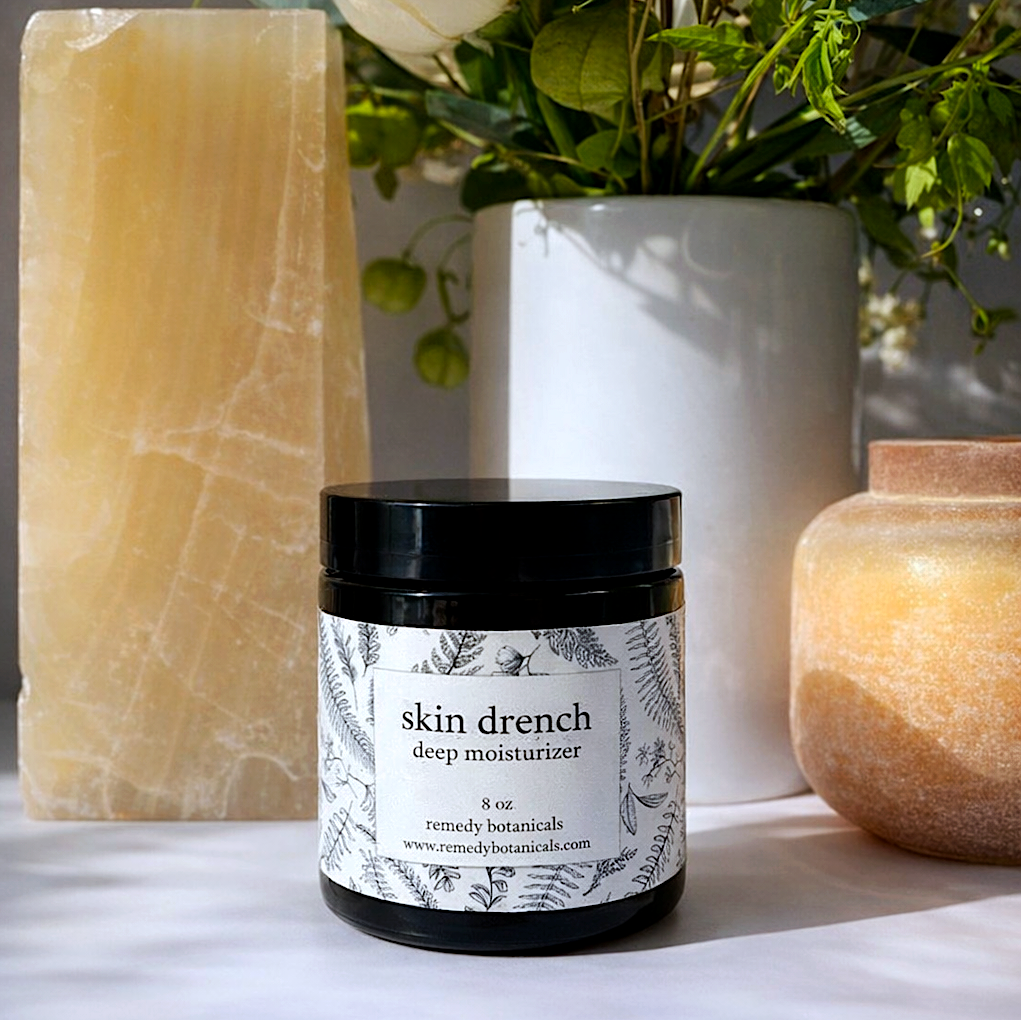 Jar of 'skin drench deep moisturizer' with a candle and flowers in the background