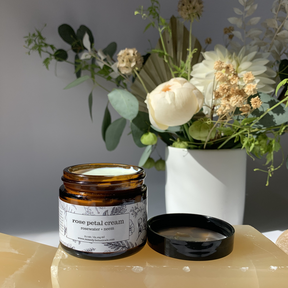 A bottle of Rose Cream stands on bright orange bars, a various florals against a soft gray wall.
