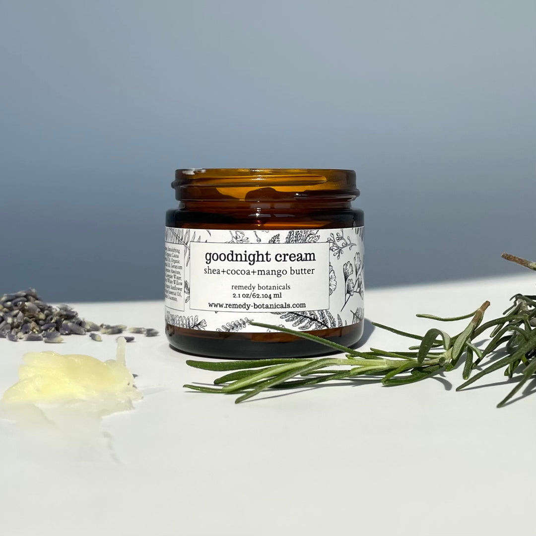 Goodnight Cream in an amber jar on a light surface, with lavender buds, rosemary, and a dab of balm.